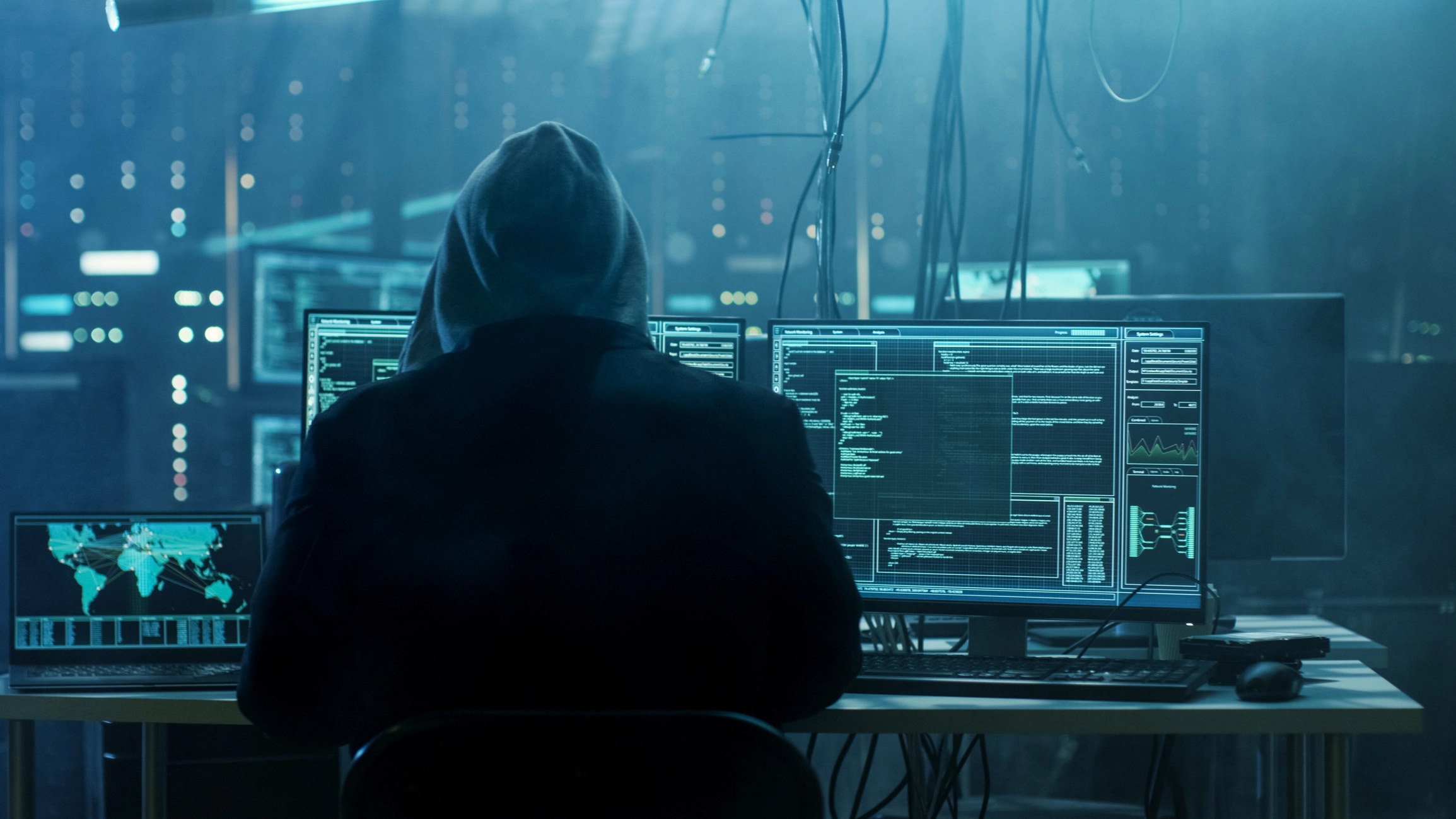 A hooded hacker sits in front of several computer monitors at a workstation inside a dark room full of data servers and cables.
