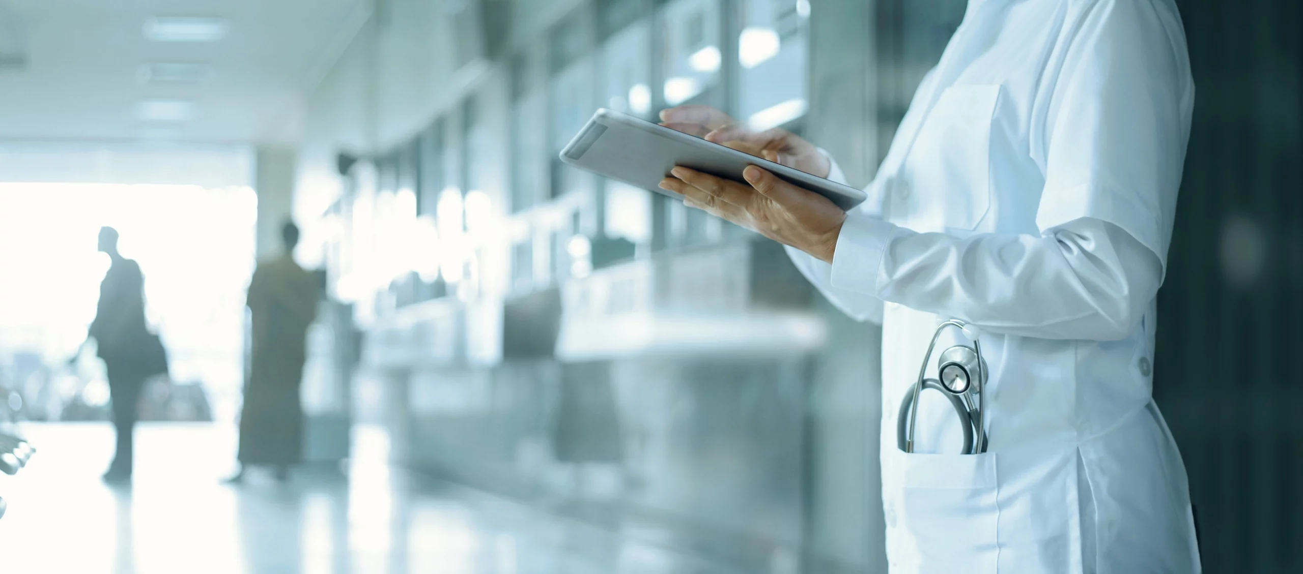 A physician checks patient information on their tablet while standing in the hallway of a hospital.