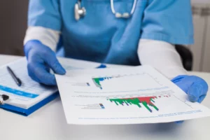A healthcare professional wearing scrubs, gloves, and a stethoscope reviews graphs of medical data on paper.