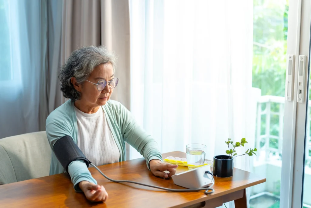 An elderly woman sitting at a table uses a blood pressure cuff.